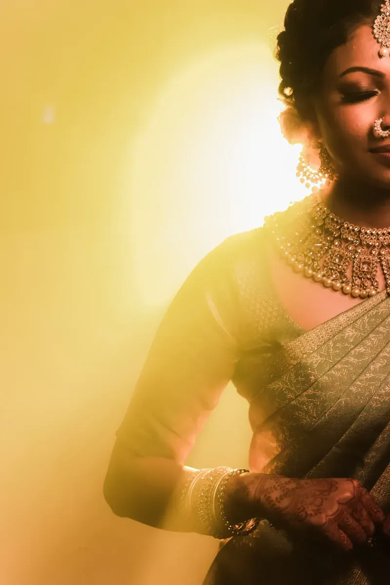 A bride in a green silk sari, her jewellery lit by warm backlight, eyes lowered.
